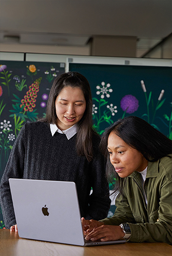 سيدتان من أعضاء فريق التسويق في Apple تعملان معاً على جهاز MacBook أمام ملصقات من تصميم فريقهما.