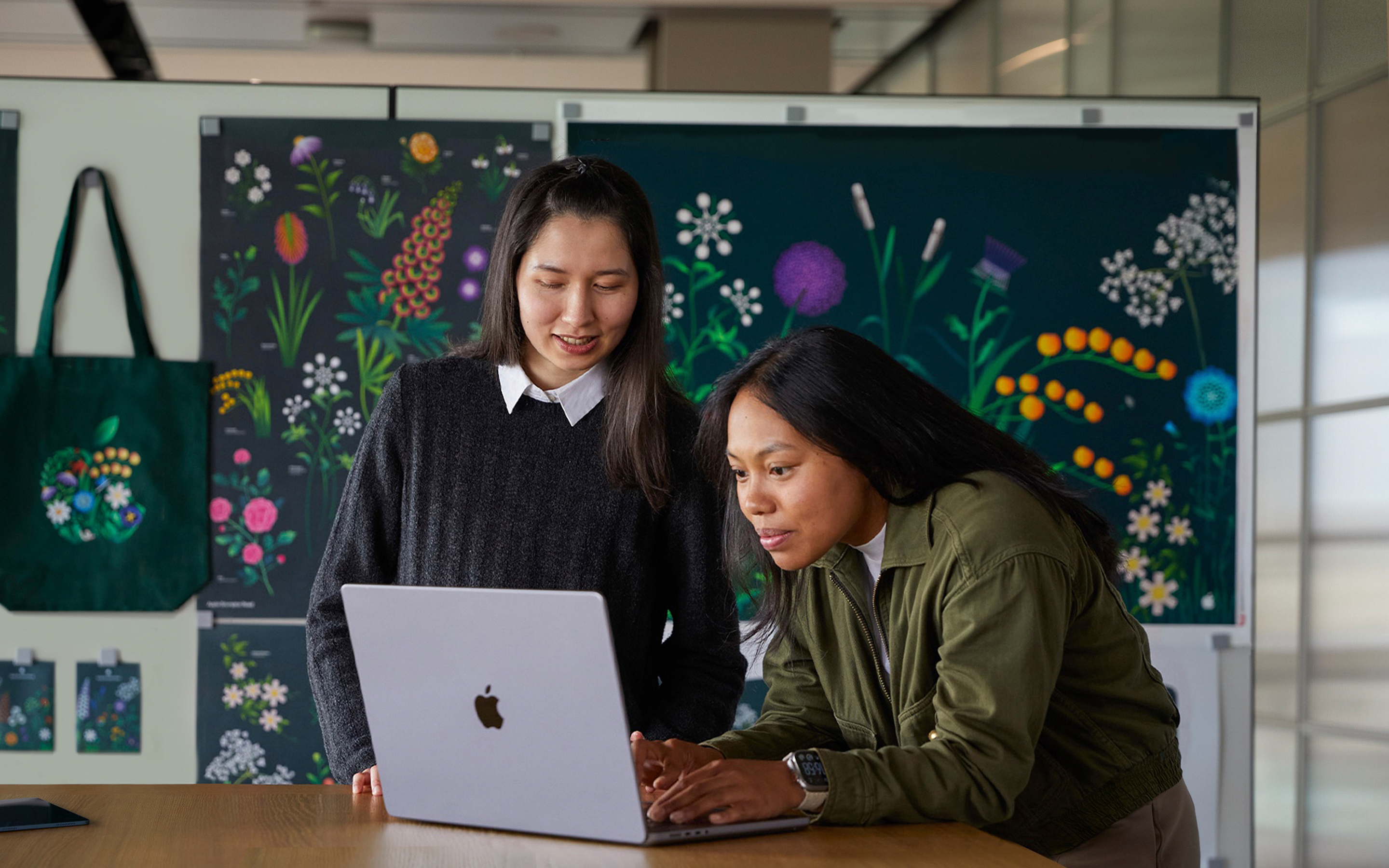 سيدتان من أعضاء فريق التسويق في Apple تعملان معاً على جهاز MacBook أمام ملصقات من تصميم فريقهما.