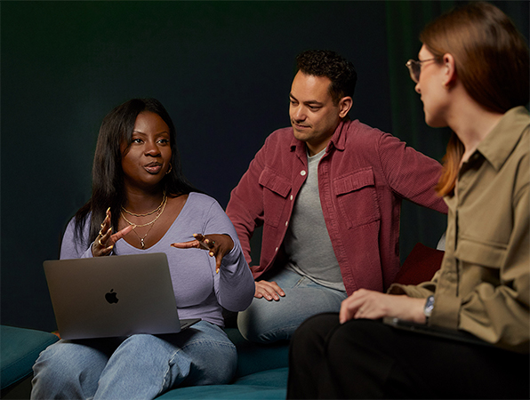 Three Apple Software and Services team members talking together on a couch, one with a MacBook.