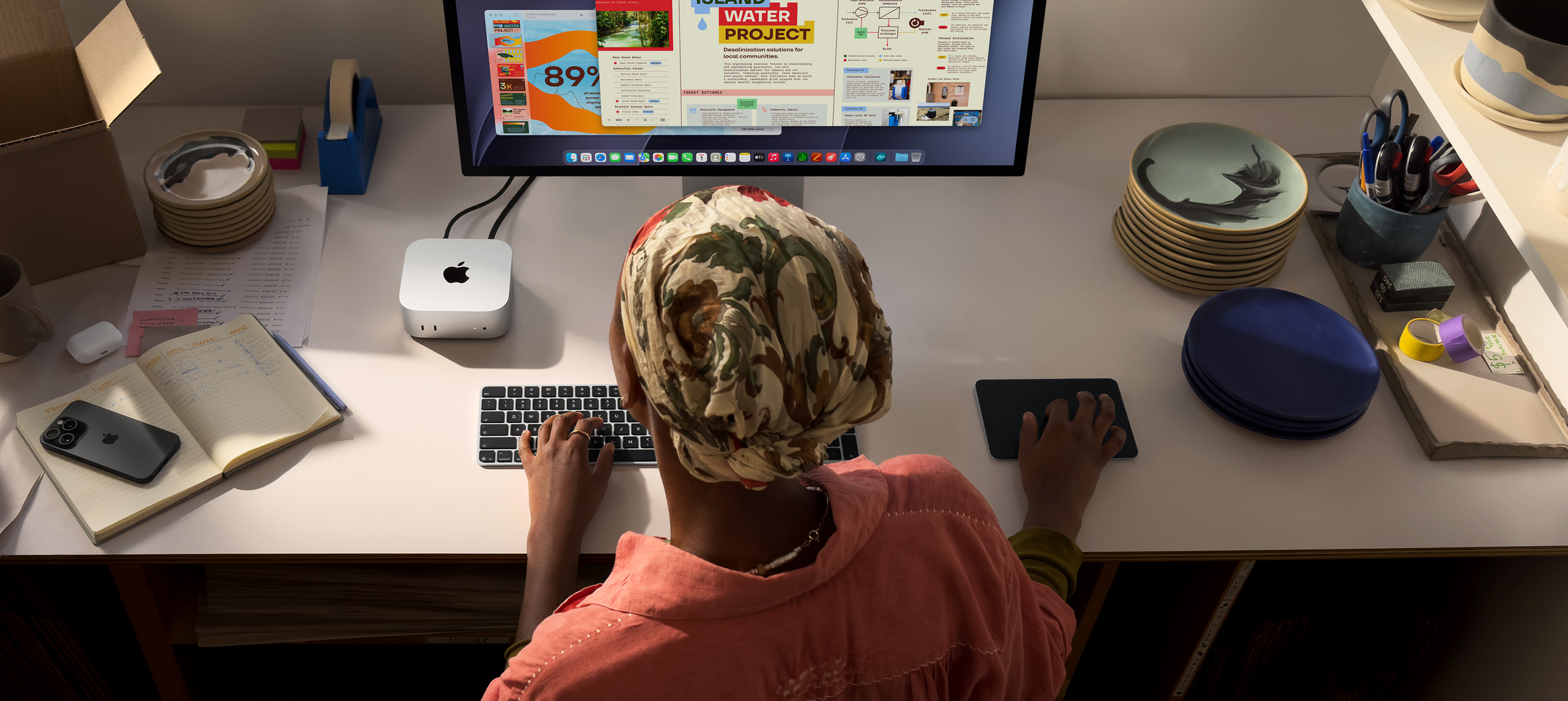 Osoba sedí u stolu a píše na Macu mini připojeném ke Studio Displayi, má Magic Keyboard, Magic Trackpad a různé další věci kolem