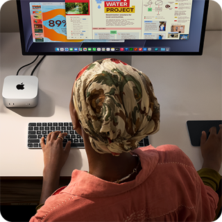 Inimene kirjutab laua taga, ees Mac mini, mis on ühendatud Studio Displayga, Magic Keyboard ja Magic Trackpad ning mitmesugused esemed