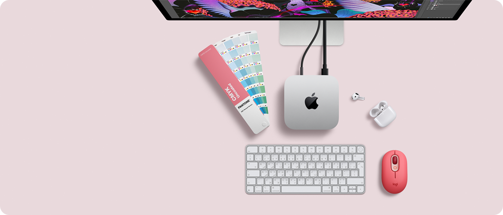 جهاز Mac mini، الجهة العلوية، ألومنيوم فضي، زوايا مدورة، شعار Apple باللون الأسود اللامع في الوسط، متصل بسلك، شاشة Studio Display تظهر بشكل جزئي في الأعلى، سماعات AirPods 4، مفتوحة، عينات ألوان Pantone، ولوحة مفاتيح رقمية وماوس