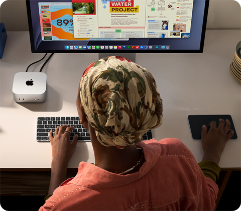 A person typing at a desk with a Mac mini, Studio Display connected, Magic Keyboard, Magic Trackpad, various items