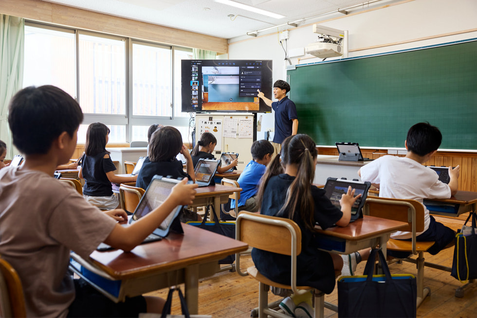 京都市の小学校の教室で前方で授業をする教師と、席でiPadを使いながらそれを見ている子どもたち。