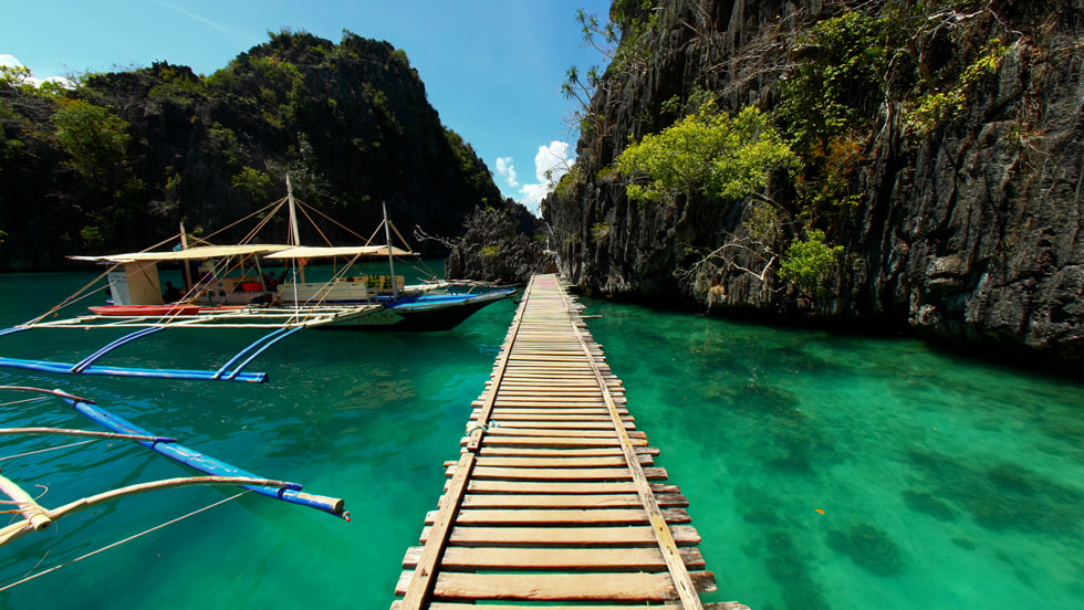 A still taken on Apple Vision Pro of an Explore POV vista that shows a wooden boardwalk extending over clear, turquoise water. On the left, a traditional outrigger boat floats in the water. 