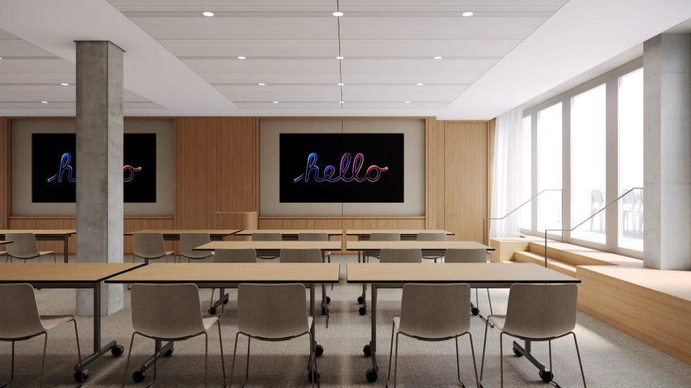 Ein leerer Schulungsraum ist mit Tischen und Stühlen ausgestattet, die auf zwei große Bildschirme an einer holzvertäfelten Wand ausgerichtet sind, auf denen jeweils das Wort „hello“ in kursiver Neonschrift angezeigt wird.
