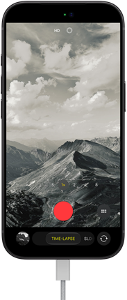 iPhone 18 Pro, вид спереду, на екрані відображається гірський пейзаж, накладене зображення режиму камери «Покадрове відео», з кабелем живлення, підключеним до зарядного порту