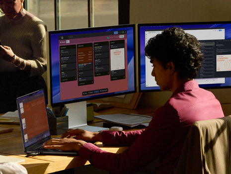 A team of employees sitting around a table in an office setting. A woman sits at her desk working on a MacBook and displays. Her colleagues are gathered around in discussion.
