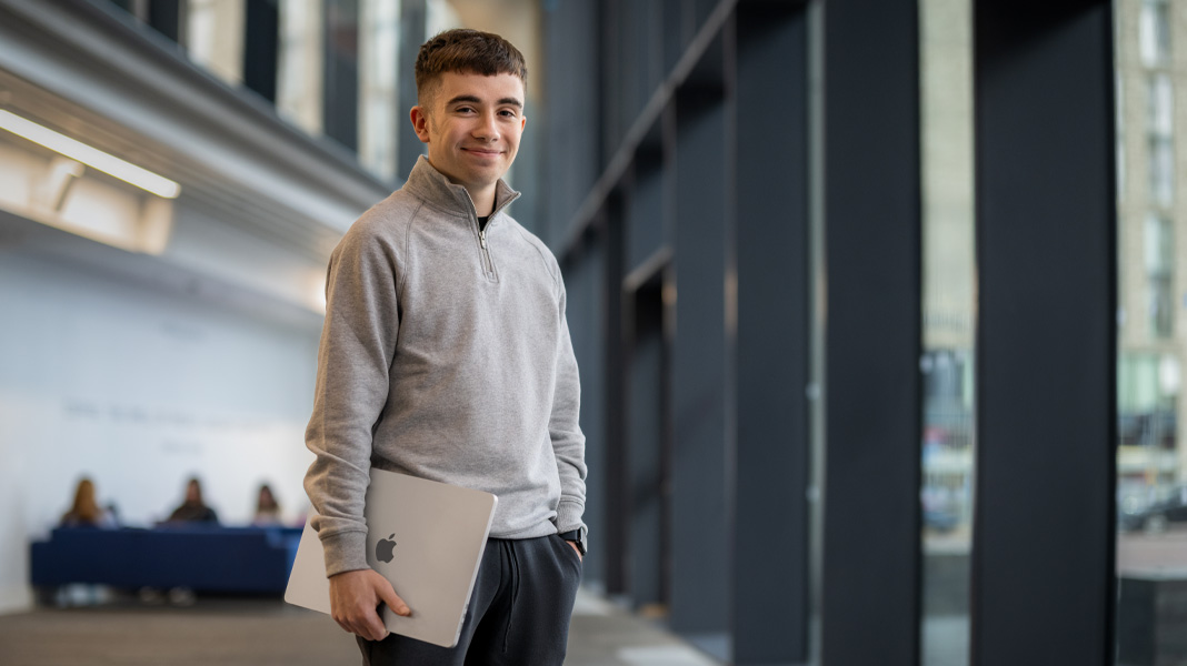 A student stands in a modern building corridor holding a MacBook laptop, smiling at the camera