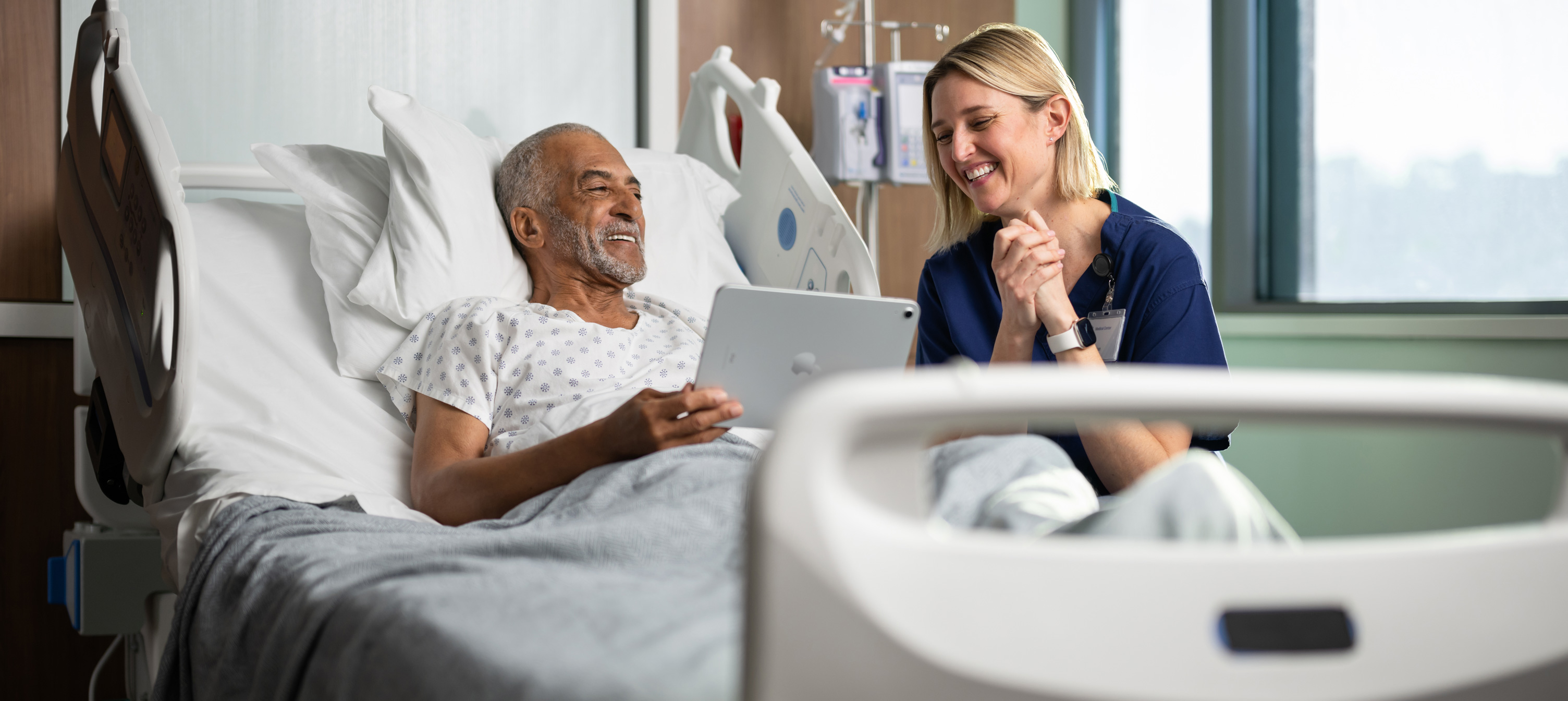 Un patient dans son lit d’hôpital regarde, tout sourire, l’écran d’un iPad avec une infirmière.