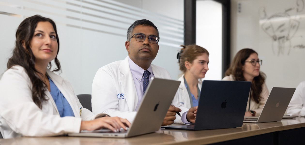 Four colleagues in doctors' coats sit in a row, each working on a MacBook.
