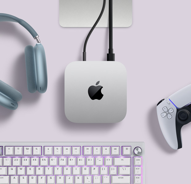 Mac mini, üstten görünüm, gümüş rengi alüminyum, yuvarlatılmış köşeler, ortalanmış parlak siyah Apple logosu, kabloyla bağlı, Studio Display standının kısmi görünümü, AirPods Max 2, mavi renk, PlayStation DualSense oyun kumandası, beyaz renk, beyaz klavye